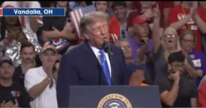 The image shows a political rally in Vandalia, OH, with a speaker at a podium and a crowd of supporters cheering and clapping.