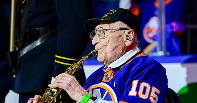 An elderly man wearing a hockey jersey and a cap is playing a saxophone at a sports event.