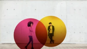 Two individuals stand in front of a wall with large, colorful circles, one in pink and the other in yellow, creating a vibrant and artistic scene.
