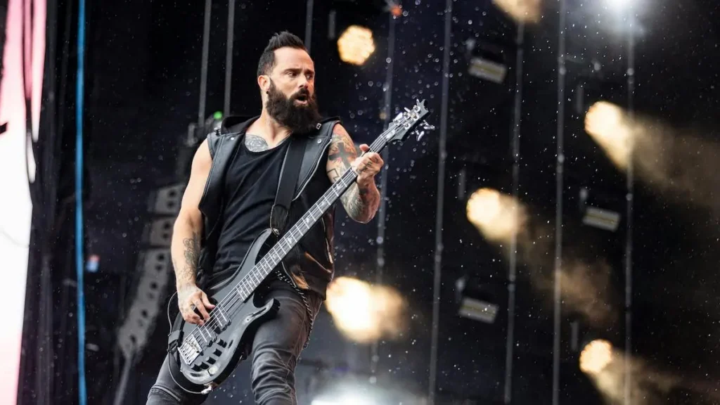 A musician with a beard and tattoos performs on stage with a bass guitar, surrounded by dynamic stage lighting.