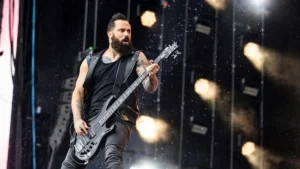 A musician with a beard and tattoos performs on stage with a bass guitar, surrounded by dynamic stage lighting.