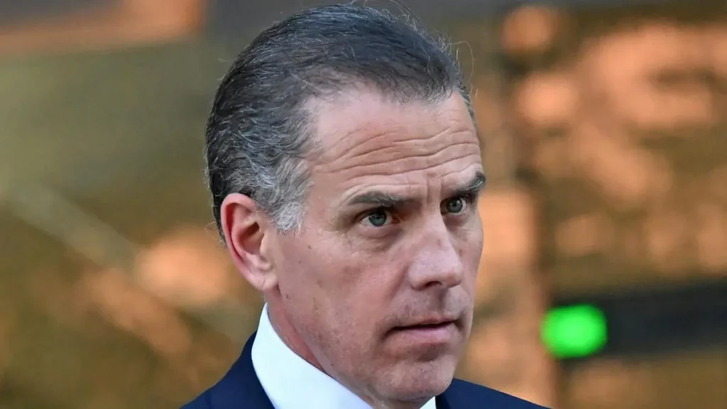 The image shows a man in a suit with a serious expression, set against a blurred background.