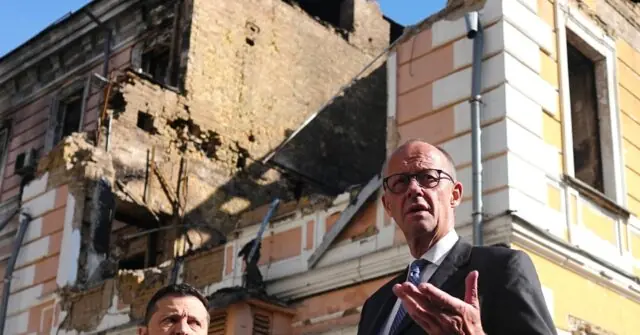 The photo shows a man in a suit speaking in front of a damaged building, possibly discussing the aftermath of an incident.