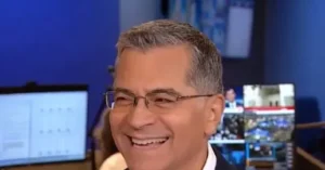 The image shows a smiling news anchor in a studio setting, with multiple screens and monitors in the background.
