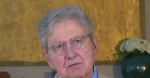 The image shows an older individual wearing glasses, seated in a room with a yellow textured wall and a decorative plant in the background.