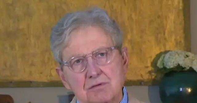 The image shows an older individual wearing glasses, seated in a room with a yellow textured wall and a decorative plant in the background.