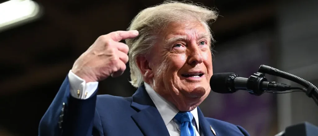 The image shows a person in a suit and tie, gesturing with their hand while speaking into a microphone.