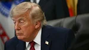 The image shows a person in a suit with an American flag pin, seated in a formal setting with flags in the background.