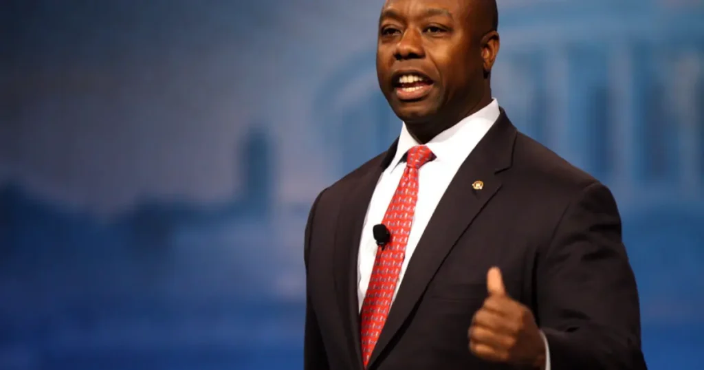 A man in a suit and tie gives a thumbs-up gesture while speaking on stage.