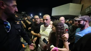 A group of people, including a man in a "Skechers" shirt, is surrounded by security personnel and media at night.