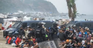 Police officers in riot gear stand in front of a crowd of people near a makeshift camp by the sea.