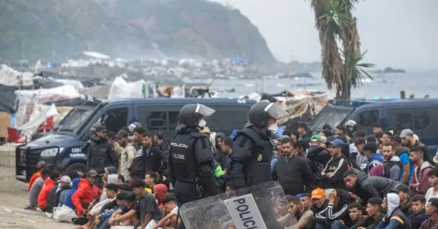 Police officers in riot gear stand in front of a crowd of people near a makeshift camp by the sea.