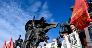 The photo depicts a group of people holding red flags and portraits, standing in front of a large statue, possibly during a commemorative or political event.