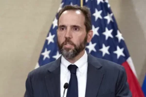 The image shows a man in a suit and tie speaking at a podium with an American flag in the background.