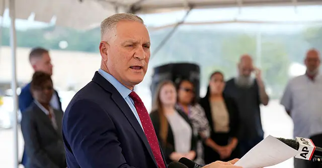 A man in a suit and tie is speaking at a press conference, with reporters and onlookers in the background.