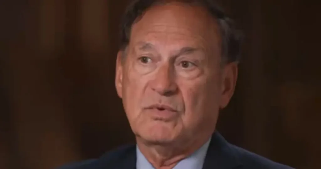 The image shows a man in a suit, speaking in what appears to be a formal or professional setting.