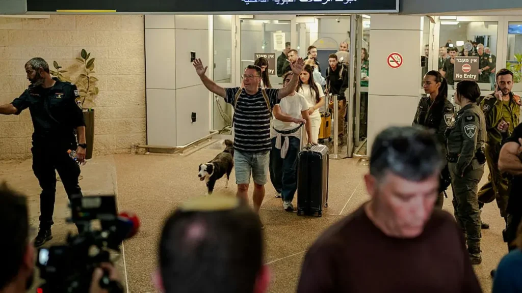 A man waves as he exits a building, accompanied by a dog, with security personnel and onlookers present.