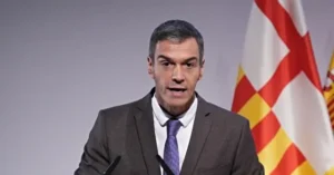 The image shows a man in a suit and tie speaking at a podium with a Catalan flag in the background.