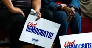 The photo shows a group of people sitting together, holding signs that say "Elect Democrats!" with the logo of the Democratic Party.