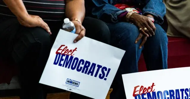 The photo shows a group of people sitting together, holding signs that say "Elect Democrats!" with the logo of the Democratic Party.
