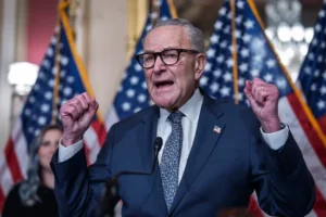 The image shows a man in a suit and tie, raising his fists in a gesture of triumph or celebration, with American flags in the background.