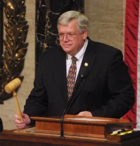 A man in a suit and tie is standing at a podium, holding a gavel, and appears to be presiding over a formal event or meeting.