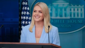 A person stands at a podium in front of a backdrop featuring the White House logo and an American flag, likely delivering a press briefing.