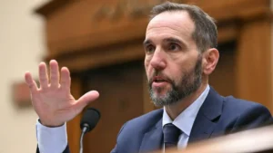 The image shows a man in a suit gesturing with his hand while speaking, likely during a formal or professional setting.