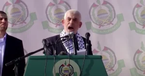 A man stands at a podium with microphones, speaking in front of a backdrop featuring the emblem of the Palestinian Authority.