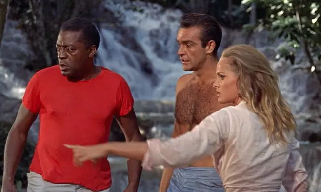 The image depicts three individuals in a natural setting with a waterfall in the background, one of whom is pointing towards something off-screen.