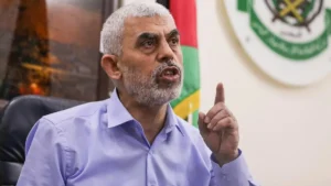 The image shows a man in a light blue shirt gesturing with his right hand, with a flag and emblem in the background.
