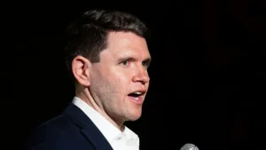 The image shows a man speaking into a microphone, dressed in a suit, against a dark background.