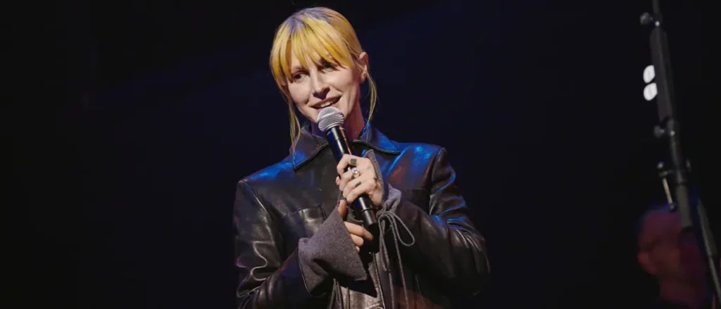 A person is holding a microphone and smiling on stage, wearing a black leather jacket.