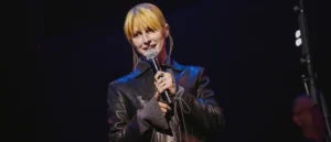 A person is holding a microphone and smiling on stage, wearing a black leather jacket.