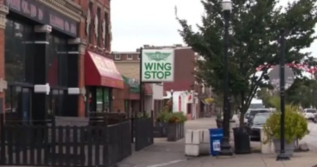 The image shows a street scene with a Wingstop restaurant sign prominently displayed on a building, surrounded by other storefronts and trees along the sidewalk.