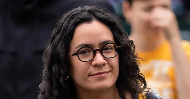 The image shows a person with curly hair and glasses, smiling slightly, with another individual in the background.