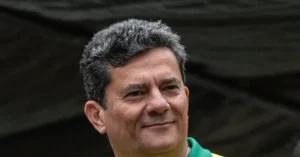 The image shows a person with short, curly gray hair, wearing a green and yellow jacket, standing in front of a dark, textured background.