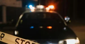 A police car with flashing lights is seen at night, with a "STOP" tape in the foreground, indicating a crime scene or emergency situation.