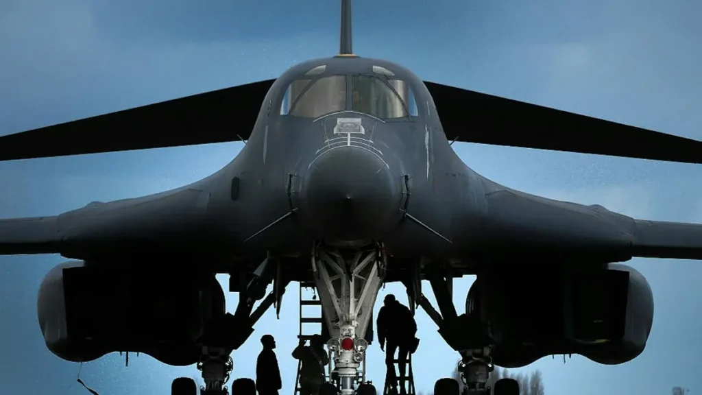 The image shows a close-up of a military aircraft with its landing gear extended, as personnel work underneath it.