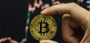 A hand holds a gold Bitcoin coin in front of a background featuring a cryptocurrency chart.