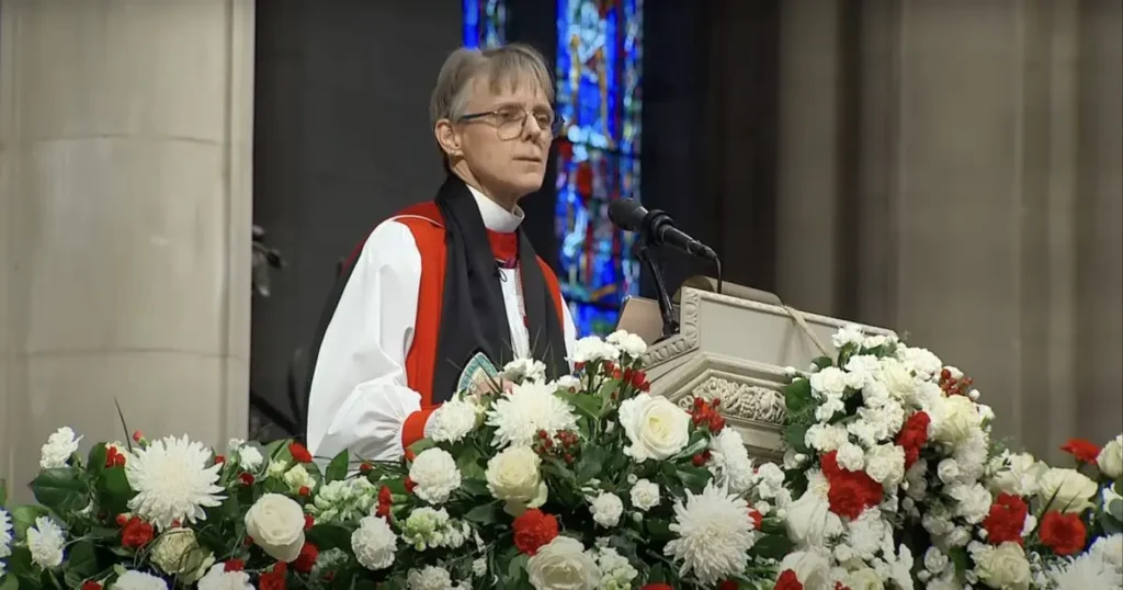 A woman in clerical attire stands at a podium adorned with flowers, delivering a speech in a church setting.
