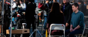 A news crew is preparing for a live broadcast, with equipment and crew members gathered around a seated reporter.