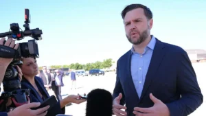 A man in a suit is giving a speech or interview outdoors, surrounded by media personnel and vehicles in the background.