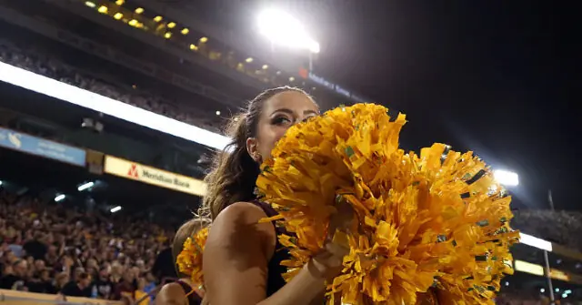A cheerleader holds large yellow pom-poms in front of a cheering crowd at a nighttime sports event.