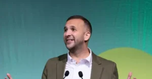 A man in a suit is speaking at a podium with a green background.