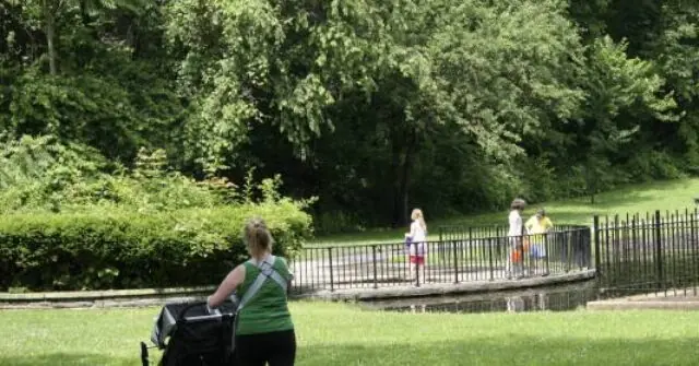 A woman pushes a stroller in a park, with children playing near a small pond and lush greenery in the background.