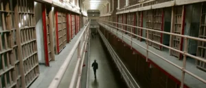 A solitary figure walks down a long, narrow corridor in a prison, flanked by rows of cells.