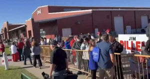 A crowd of people gathers outside a building, some wearing red hats, as they wait in line, possibly for an event or election.