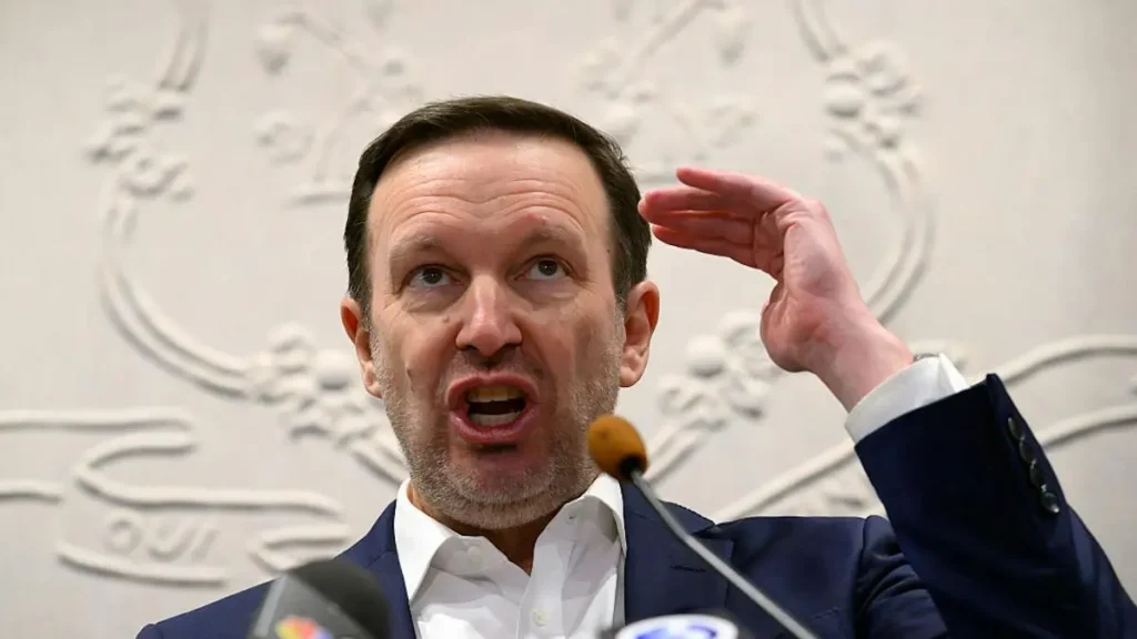 A man in a suit gestures with his hand while speaking into a microphone during a press conference.