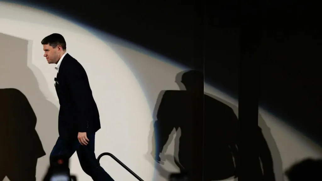 A man in a suit walks down a staircase, with his shadow cast prominently on the wall behind him.